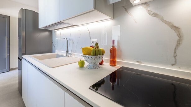 A modern kitchen with a white countertop, a black induction cooktop, a stainless steel refrigerator, and a bowl of fruit.