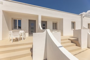 A white building with a staircase leading to a door, a table with chairs outside, and a clear blue sky. - Appartamenti Mancaversa (Taviano)
