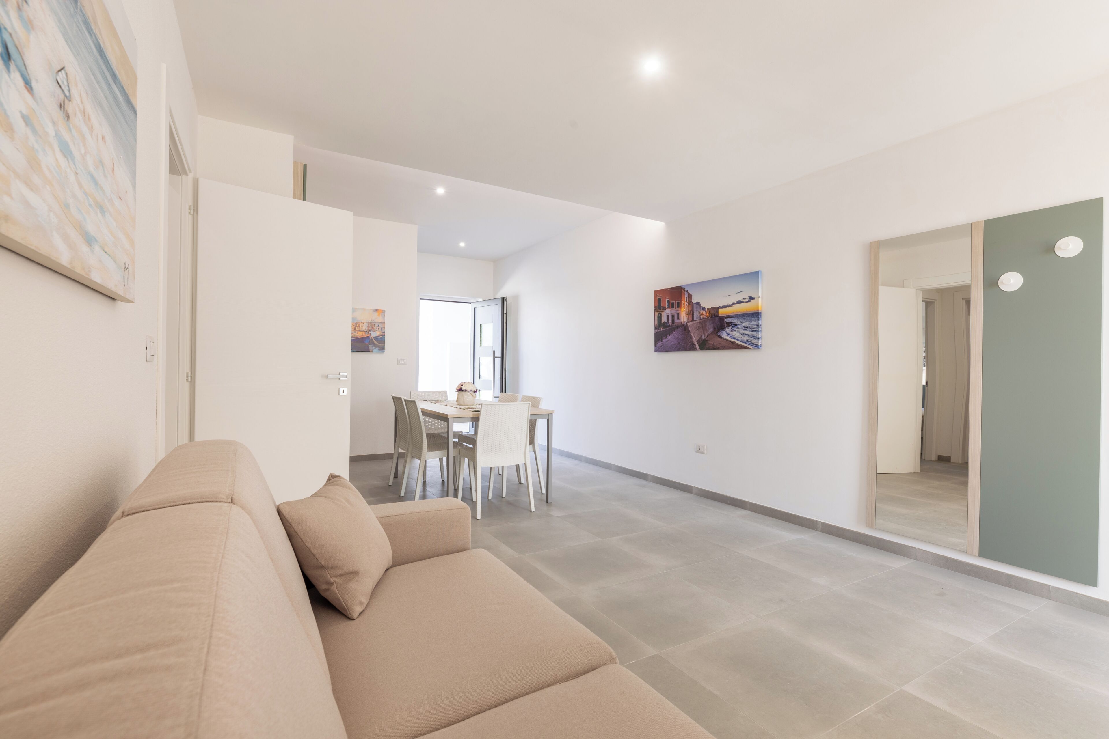 Een moderne woonkamer met een beige bank, een eethoek met stoelen en een tafel, en een grote spiegel aan de muur.