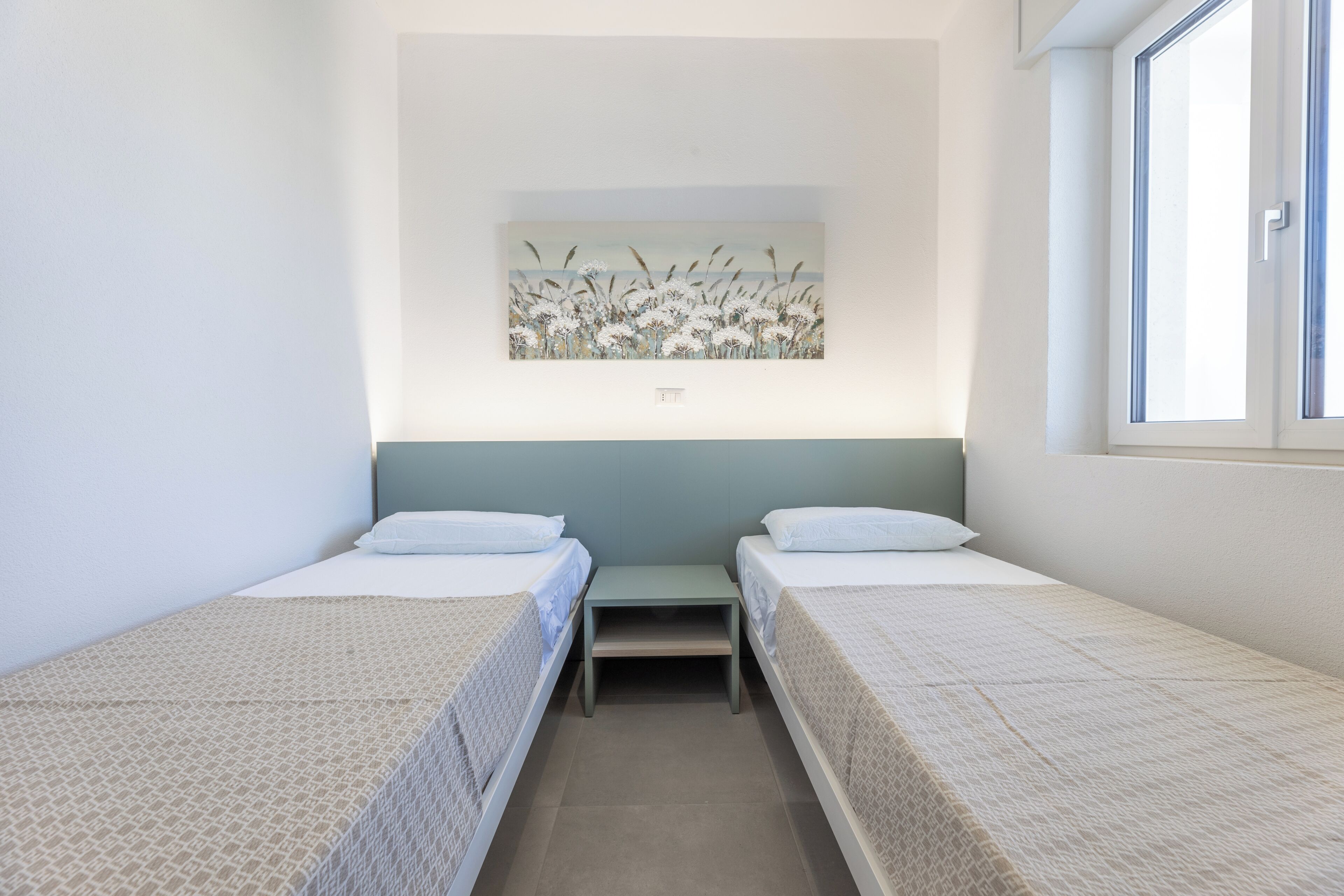 A room with two beds, a small table, a painting on the wall, and a window.