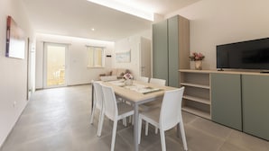 A modern kitchen with a flat-screen TV, a dining area with white chairs and a table, and a sliding door leading to a balcony.