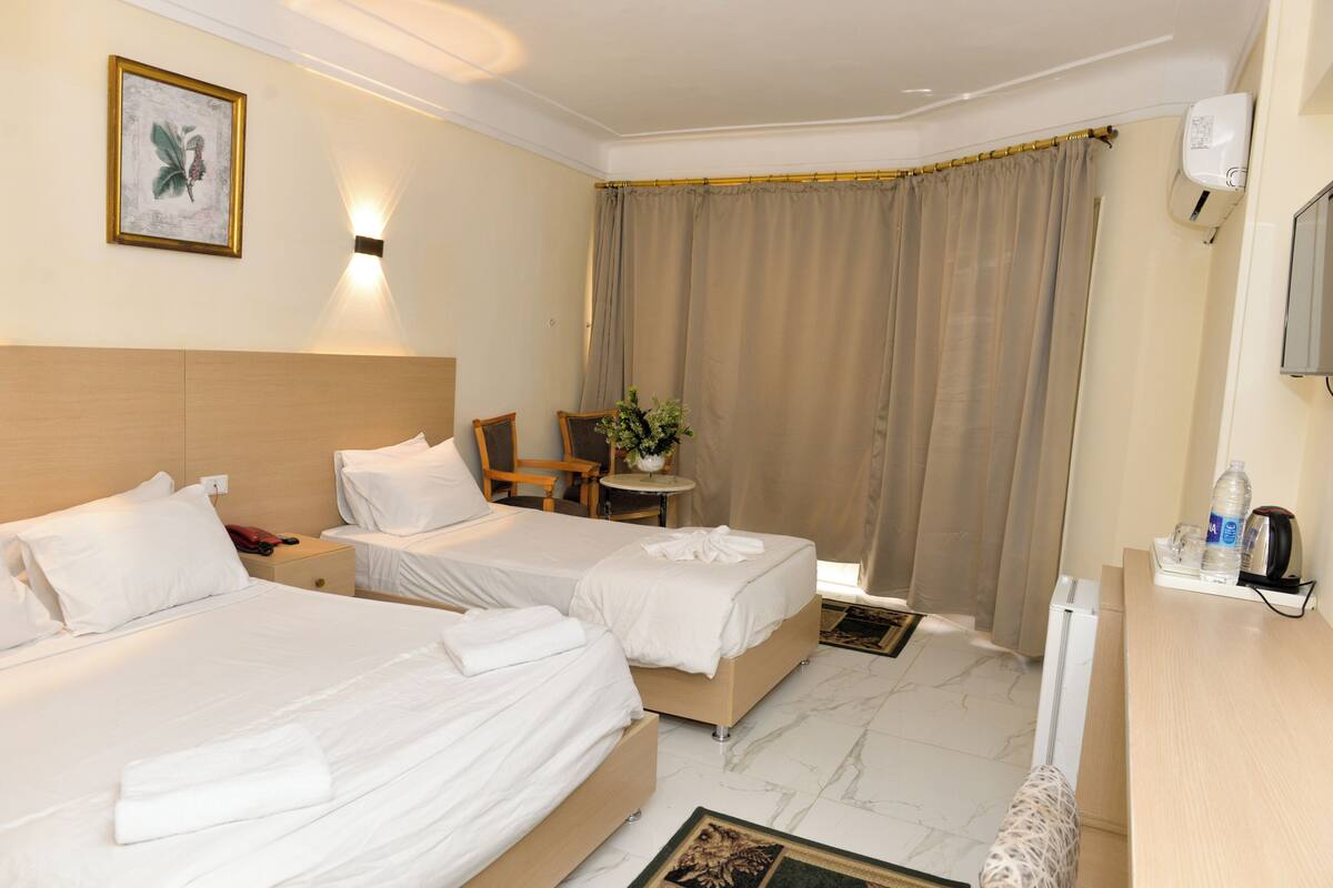A hotel room with two beds, a small table with a plant, a chair, and a desk with a water bottle and coffee maker.