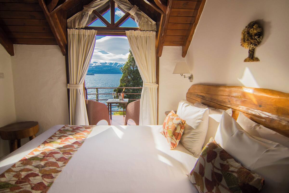 A bedroom with a large bed, a wooden headboard, and a view of a lake and mountains through a window.