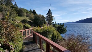 A wooden bridge with metal railings leading to a lakeside area with dense greenery and a view of distant mountains.