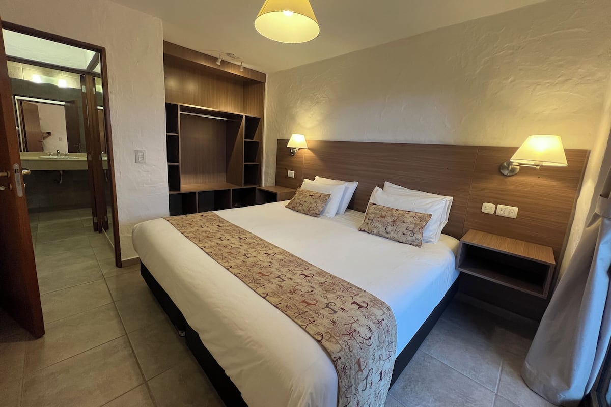 A hotel room with a large bed, bedside lamps, a wooden headboard, a built-in wardrobe, and a bathroom visible through an open door.