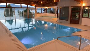 An indoor swimming pool with clear glass walls, a hot tub, and a view of the mountains.