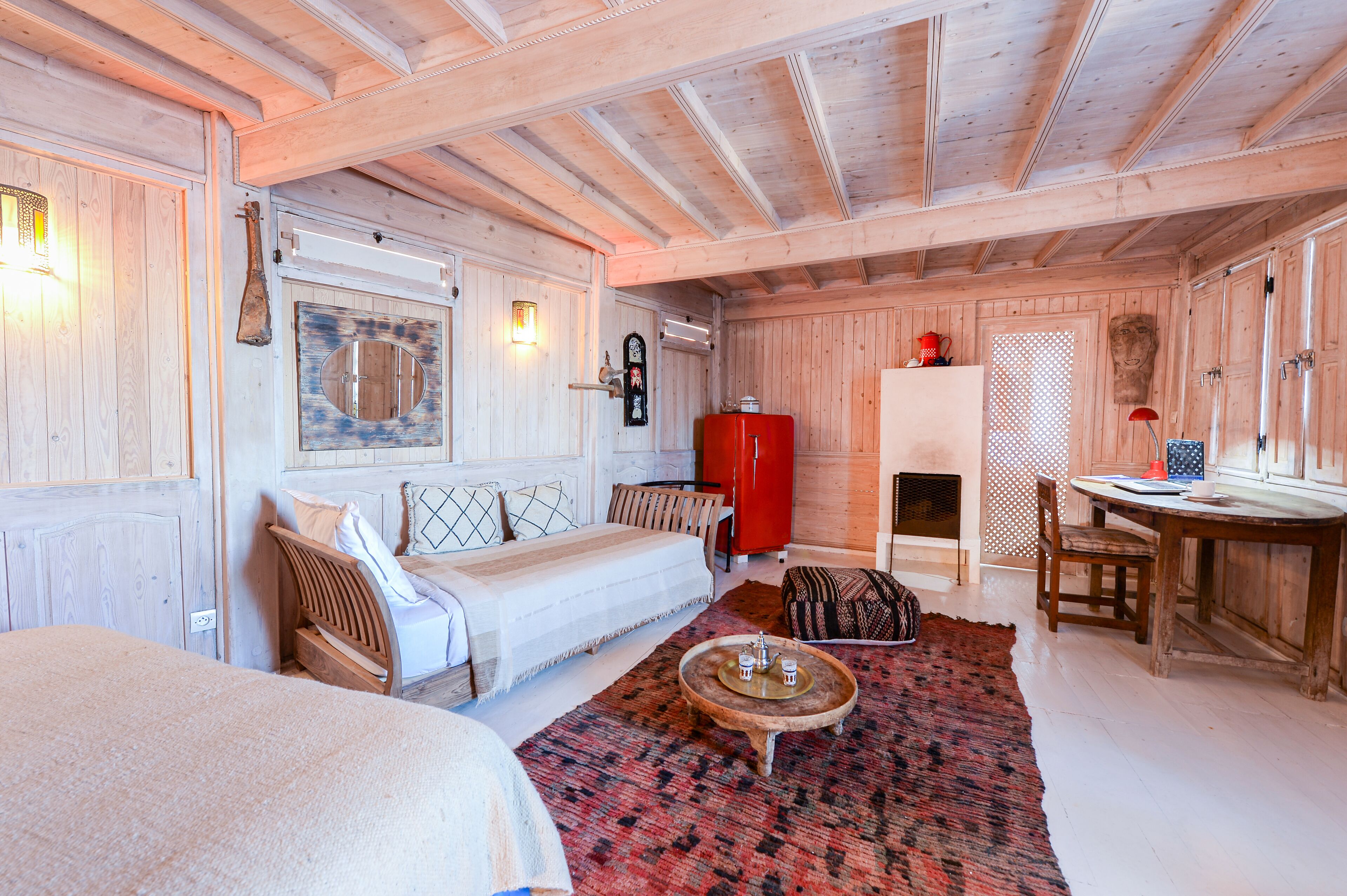 A cozy wooden cabin with a bed, a red refrigerator, a dining table, and a fireplace.