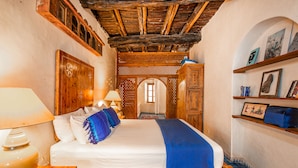 A traditional room with a wooden bed, a nightstand with a lamp, a wooden wardrobe, and a shelf with framed pictures. - Dar Cherazade & Spa (Essaouira)