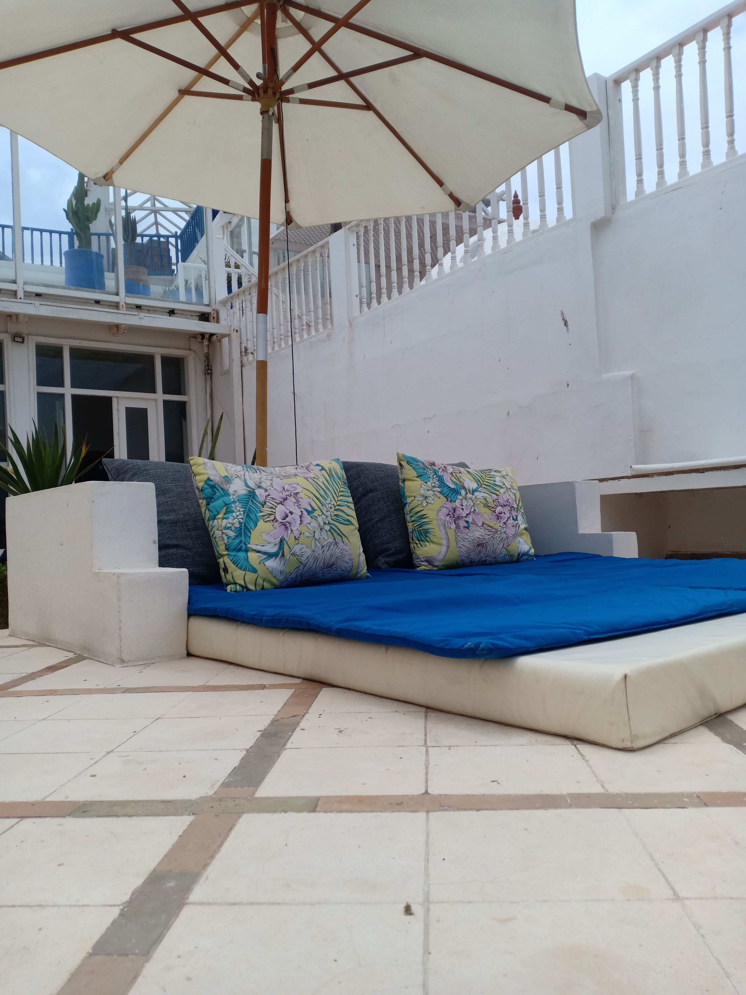 Une terrasse dotée d’un coin salon avec des coussins bleus, protégé par un grand parasol, avec des murs blancs et un sol carrelé.