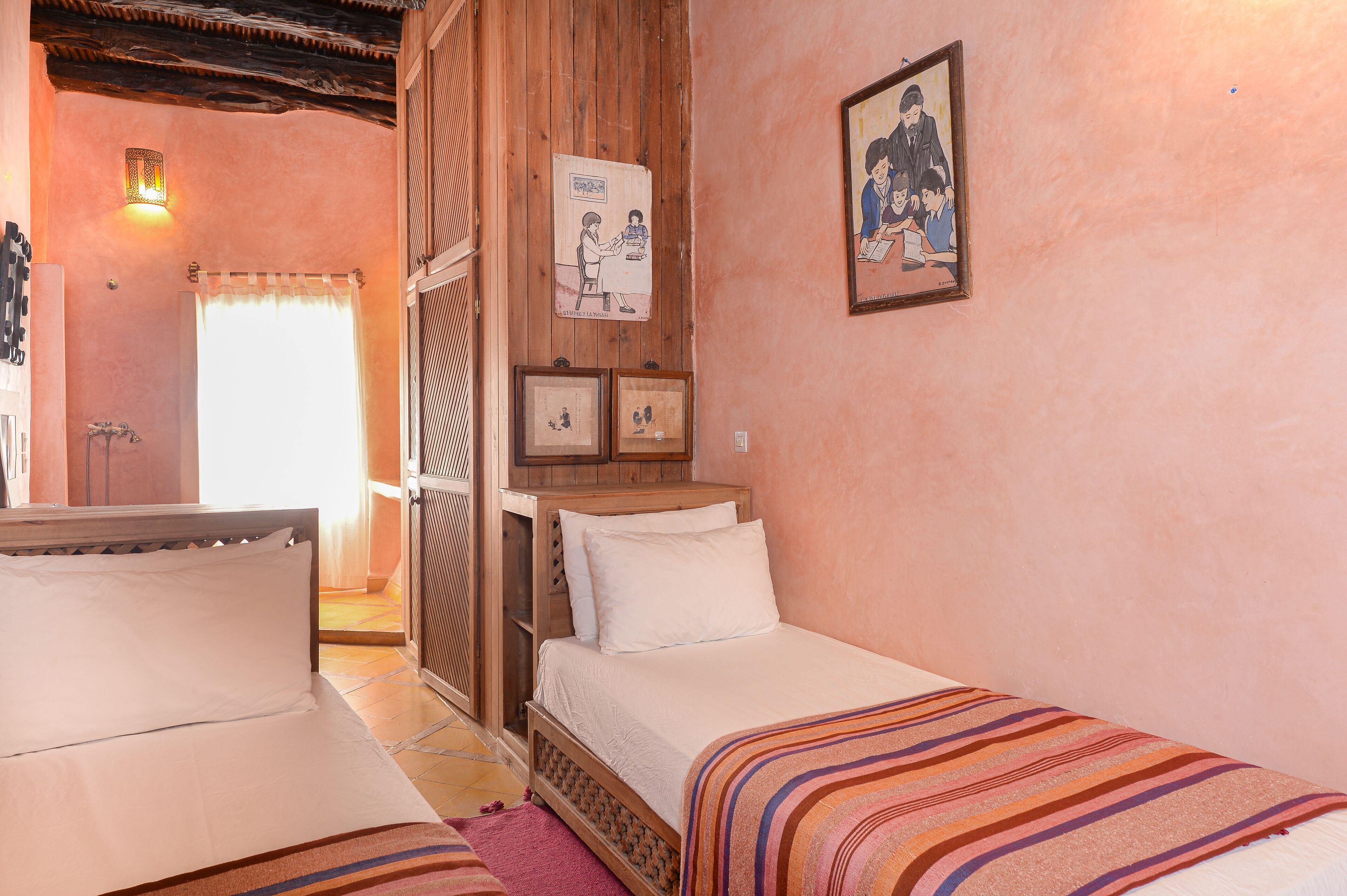 A bedroom with two beds, wooden headboards, a wooden wardrobe, and a wall-mounted shelf with framed pictures.