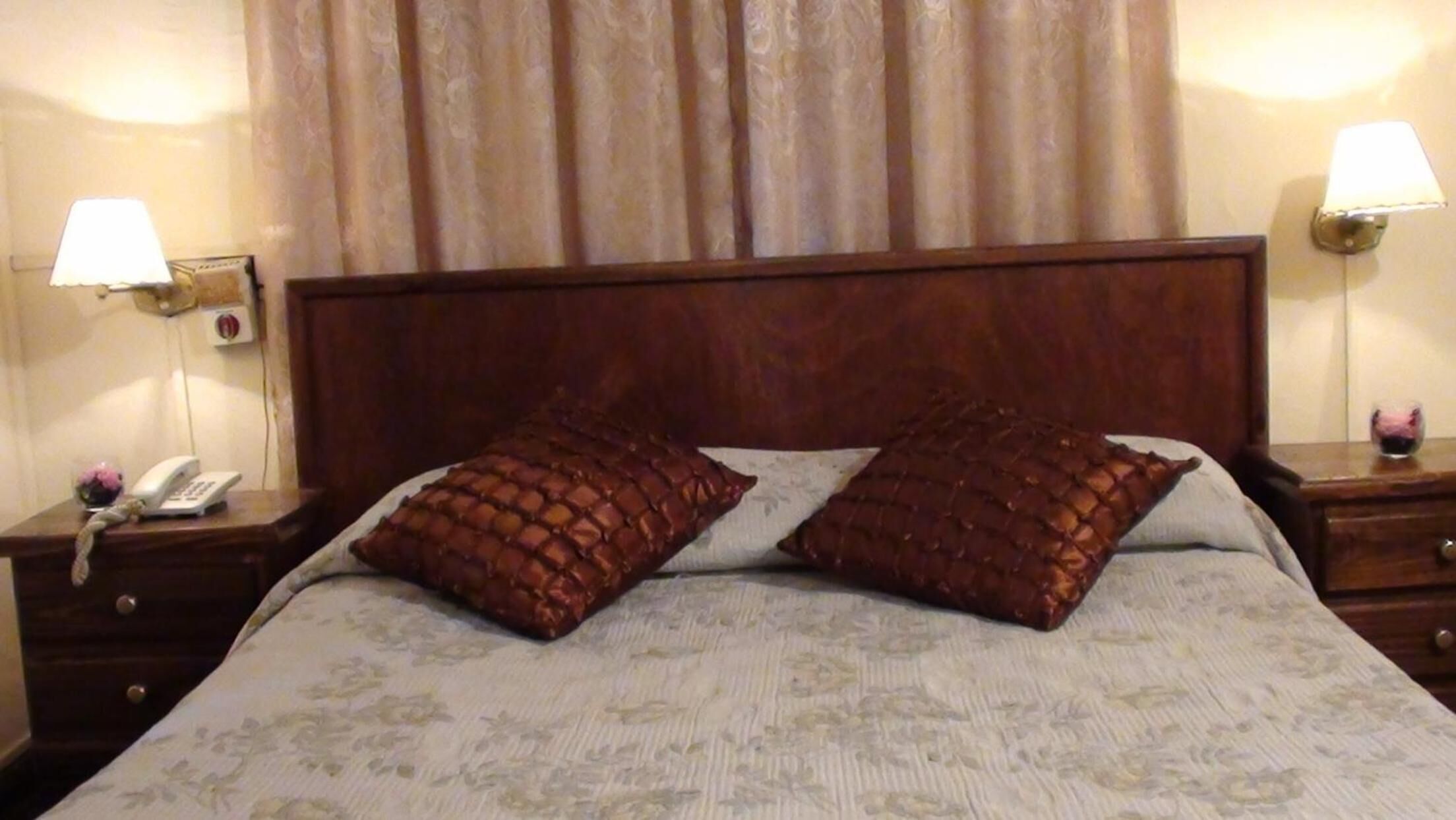 A hotel room with a wooden headboard, two bedside tables with lamps, a telephone, and two decorative pillows.