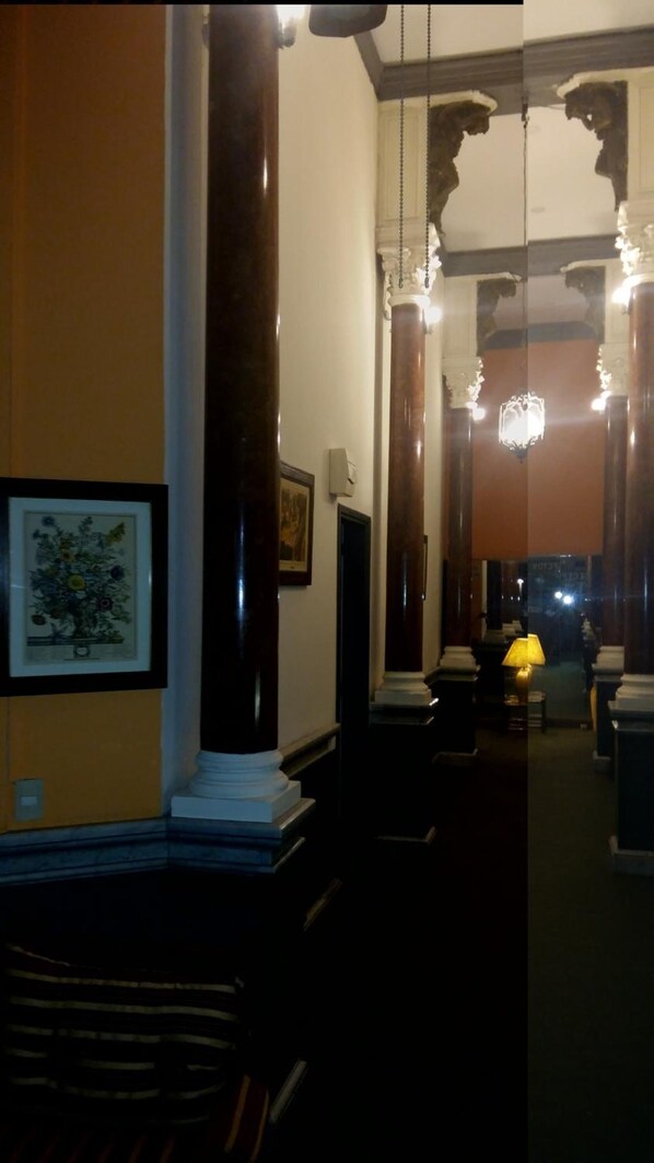 A hallway with a framed floral artwork on the left, a dark wooden column, and a chandelier in the distance.