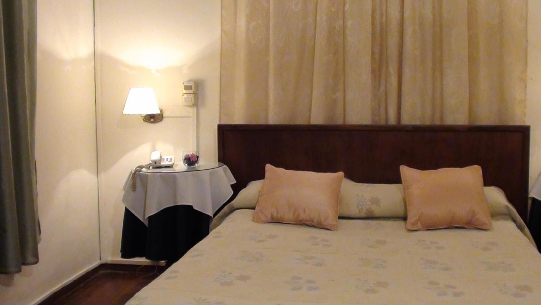 Een hotelkamer met een groot bed, twee kussens, een nachtkastje met een lamp en een wandthermostaat.