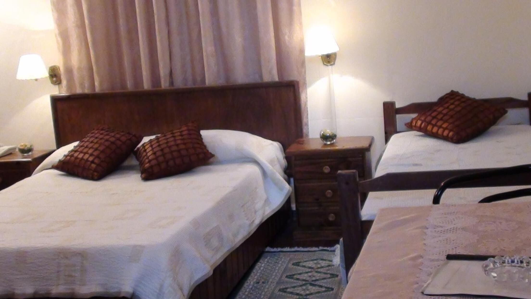 A hotel room with two beds, each with a wooden headboard and a white bedspread. There are two bedside tables with lamps, a small table with a bowl, and a patterned rug on the floor.