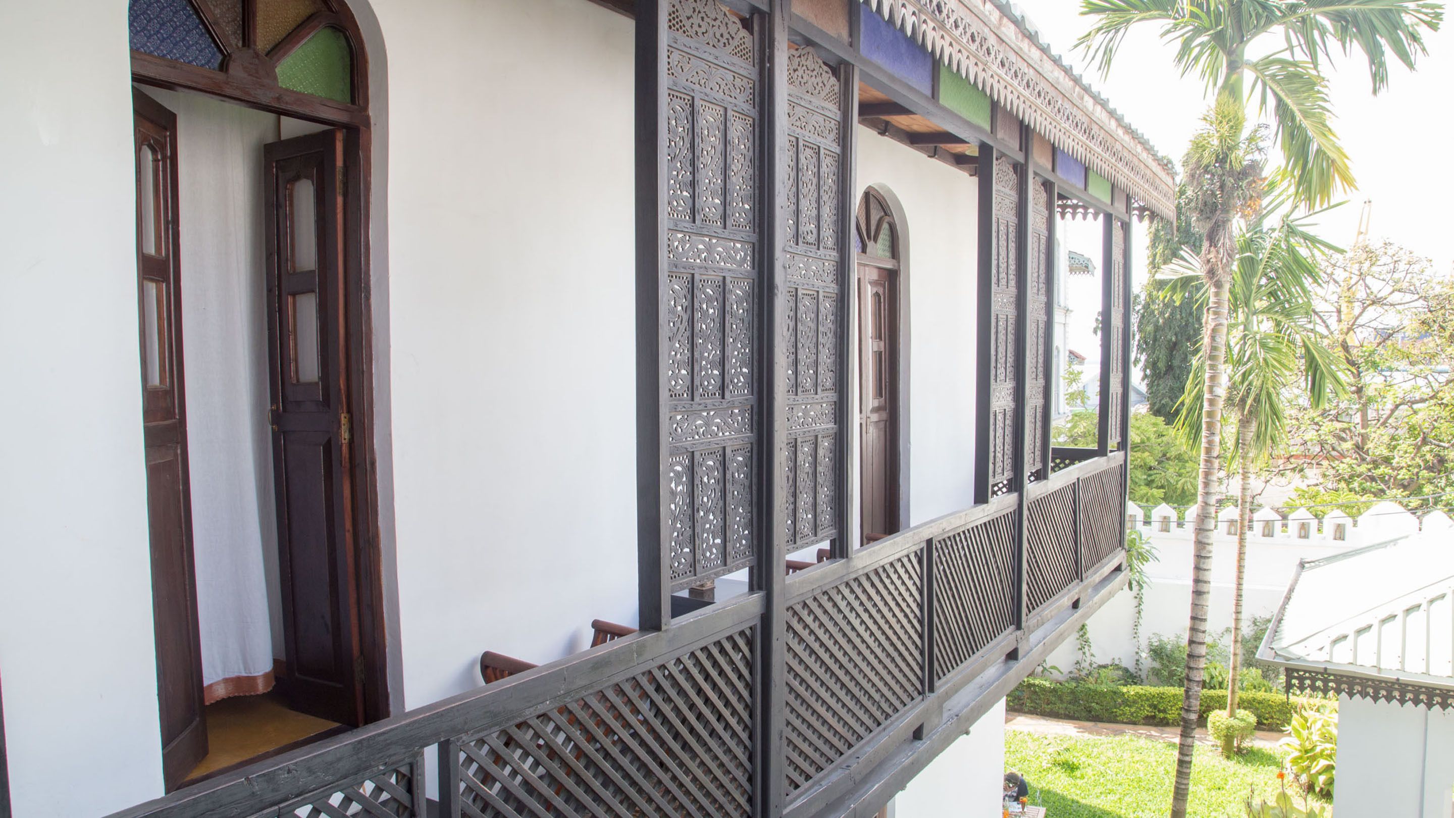 A traditional building with ornate wooden doors and windows, a balcony with lattice railings, and a view of a garden with palm trees.