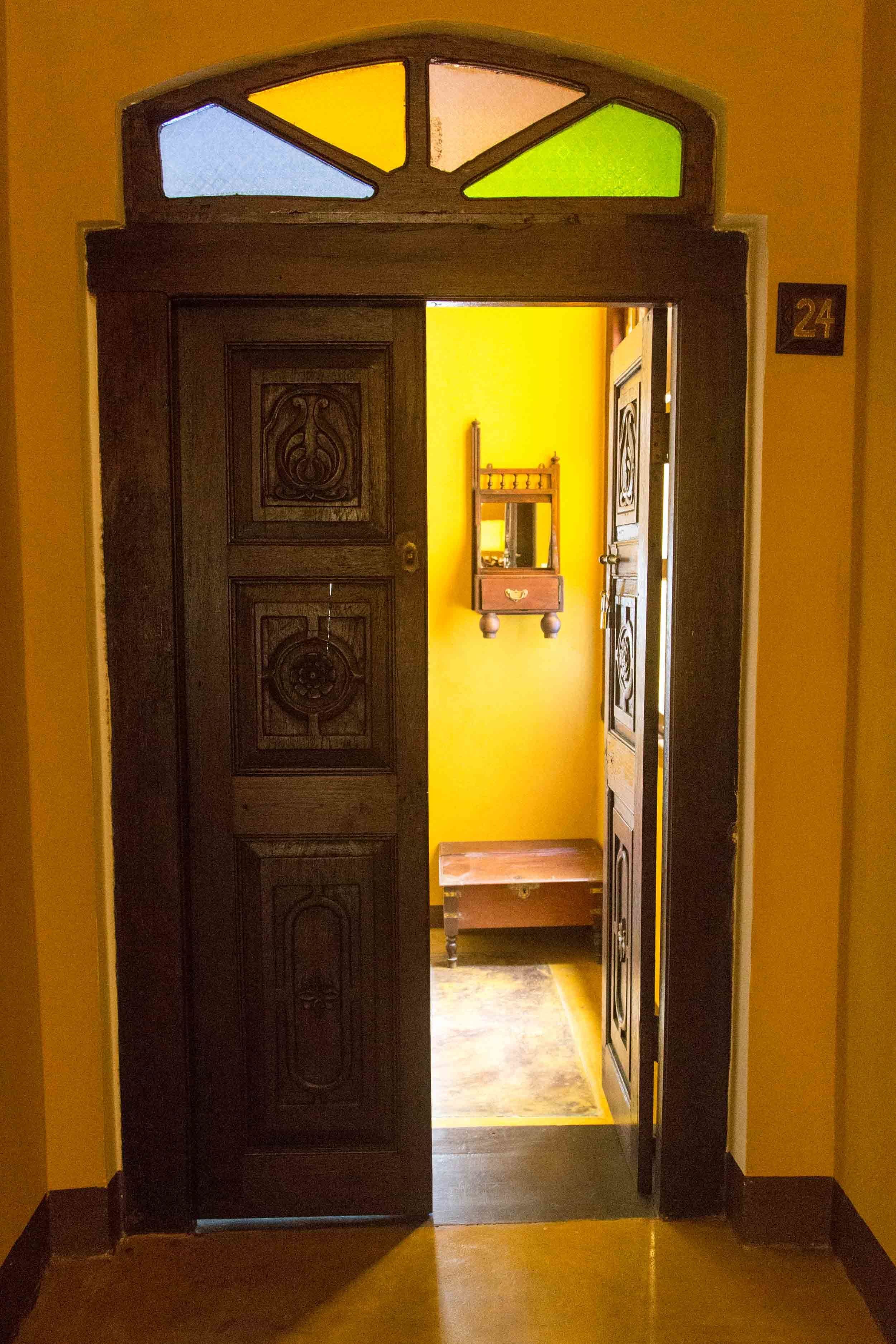A wooden door with a stained glass window above it, leading into a room with a yellow wall and a small wooden bench.