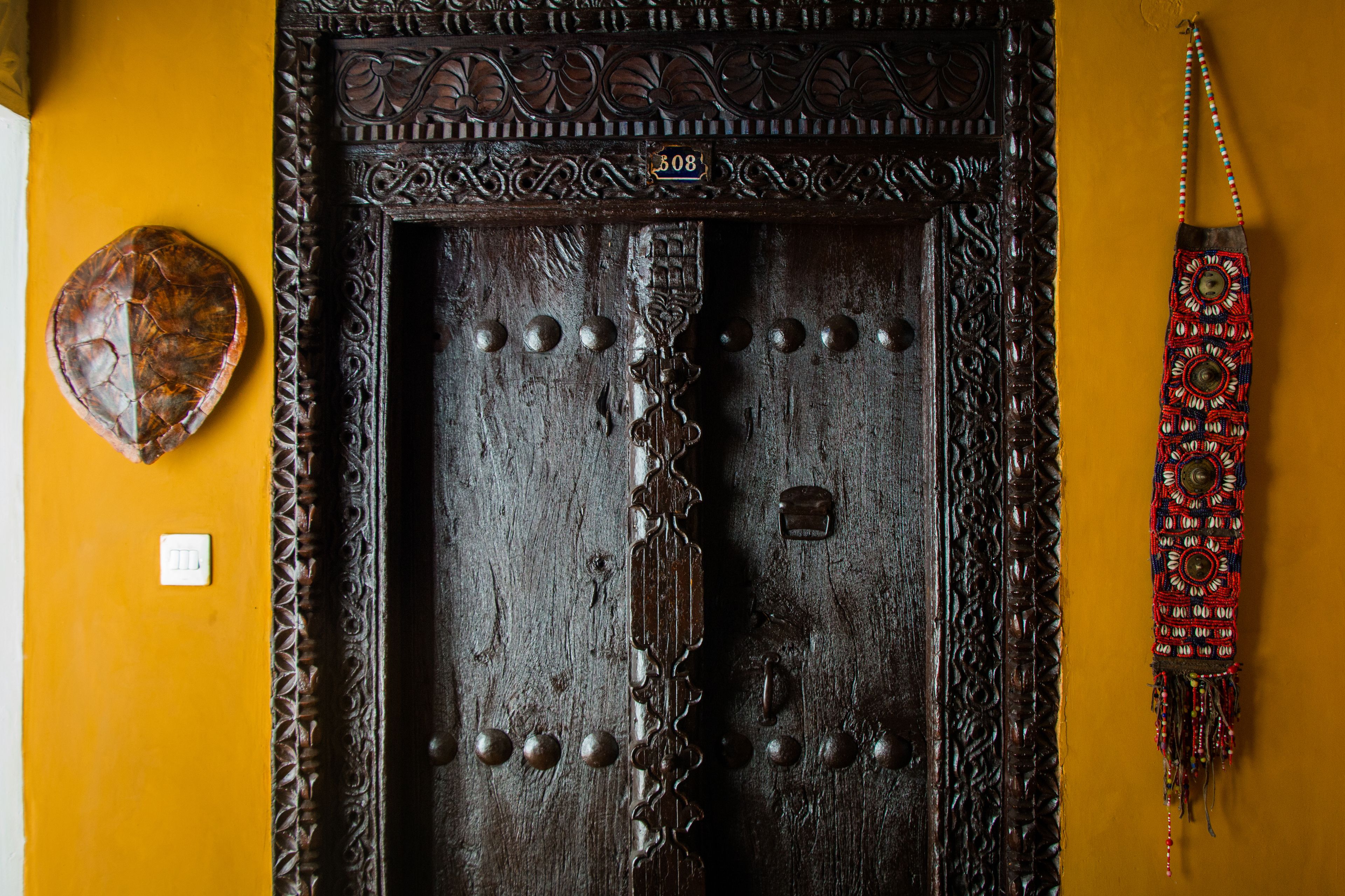 A wooden door with intricate carvings and metal studs, a decorative wall hanging, and a small white switch on a yellow wall.