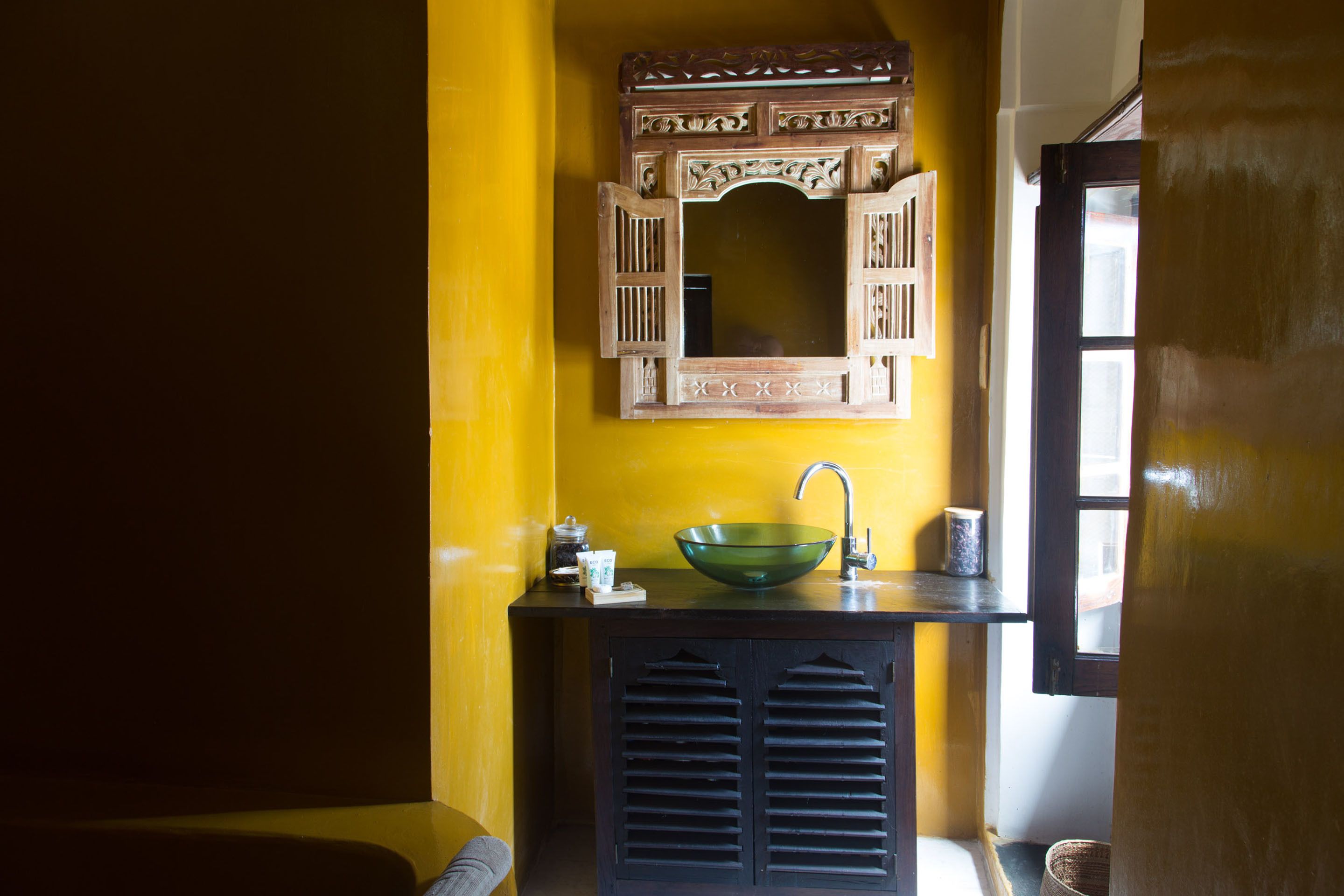 A bathroom with a yellow wall, a wooden cabinet, a green glass sink, and a mirror above it.
