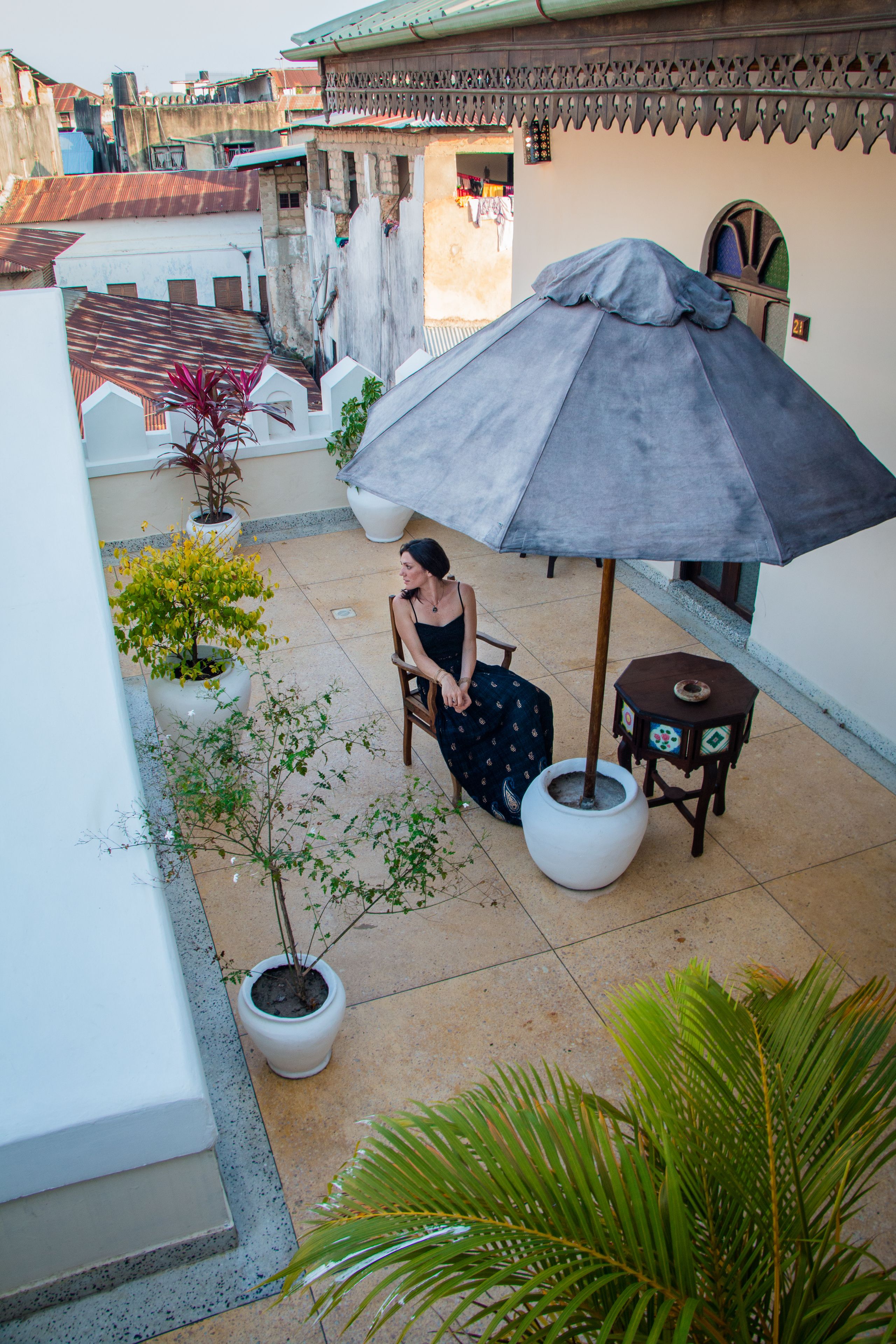 Una donna seduta sotto un ombrello grande su un balcone con piante in vaso e pavimento piastrellato.