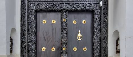 Una puerta de madera oscura con tallas intrincadas y detalles dorados, ubicada en una pared blanco y beige con un pequeño arco en la parte superior.
