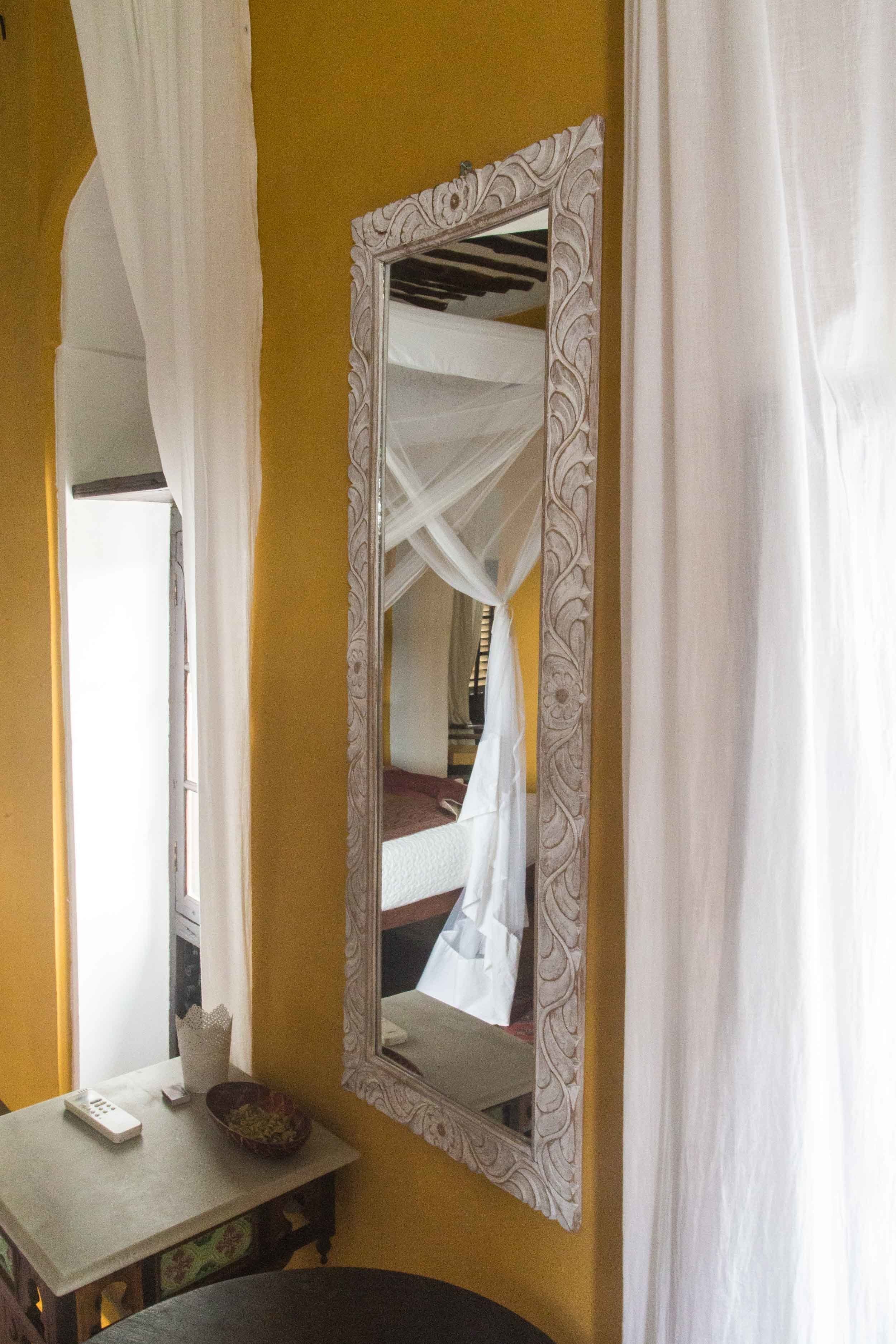 Une chambre avec un lit, une table de chevet sur laquelle se trouvent une télécommande et un bol, et un grand miroir décoratif accroché au mur.