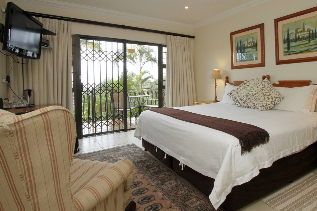 A hotel room with a bed, a chair, a nightstand, a TV, and a balcony with a view of palm trees.