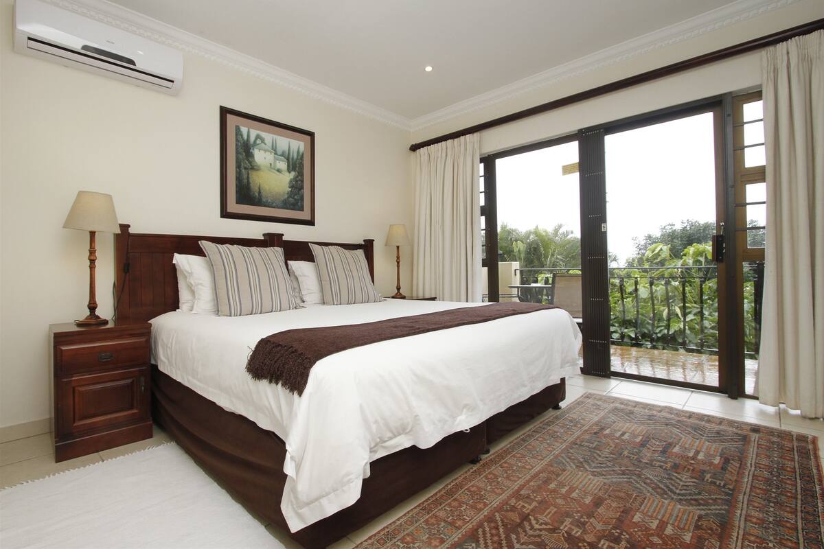 A bedroom with a large bed, a nightstand, a lamp, a painting, and a view of greenery through a sliding glass door.