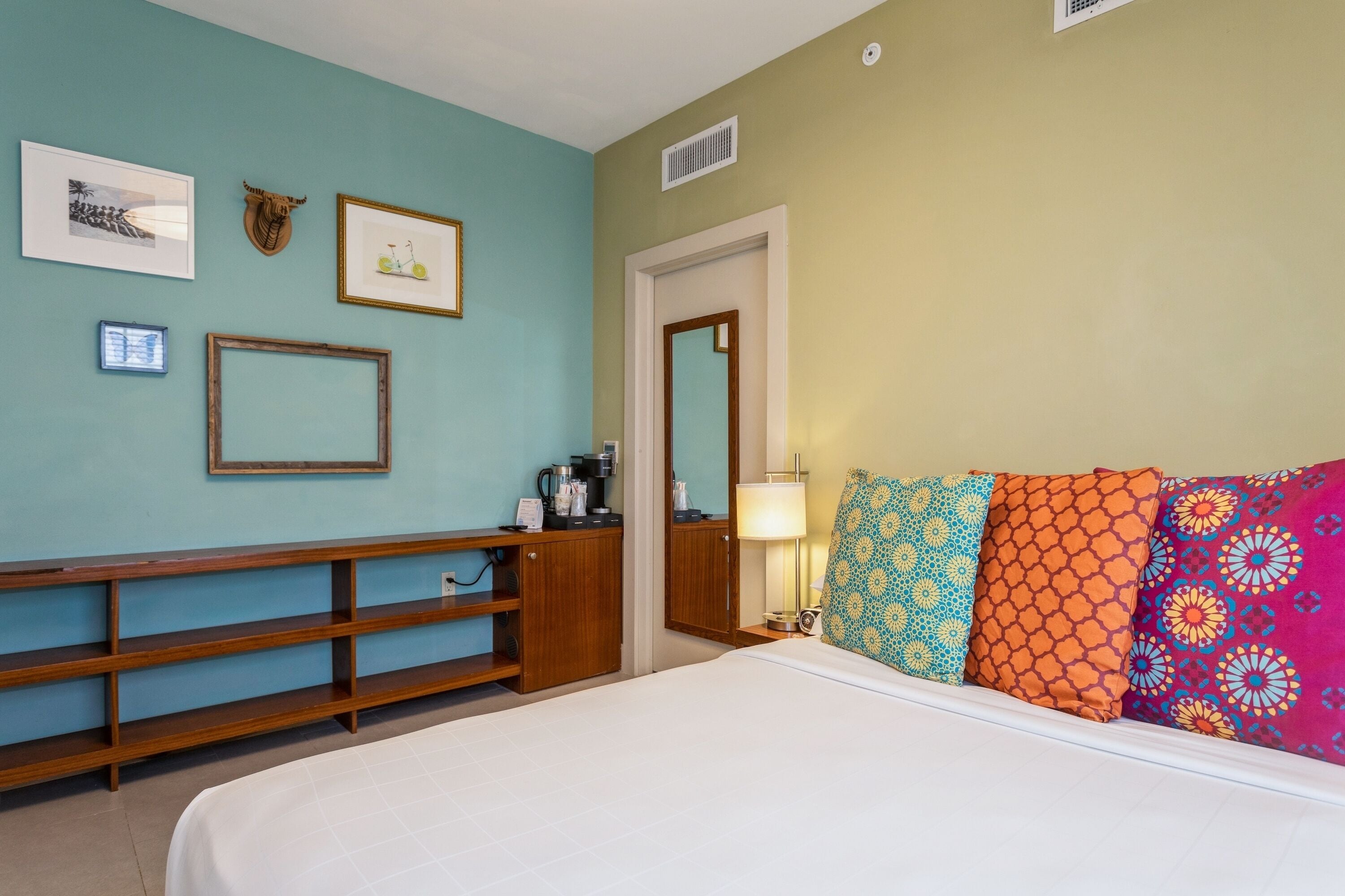 A hotel room with a bed, a wooden bench, a coffee machine, and a mirror.