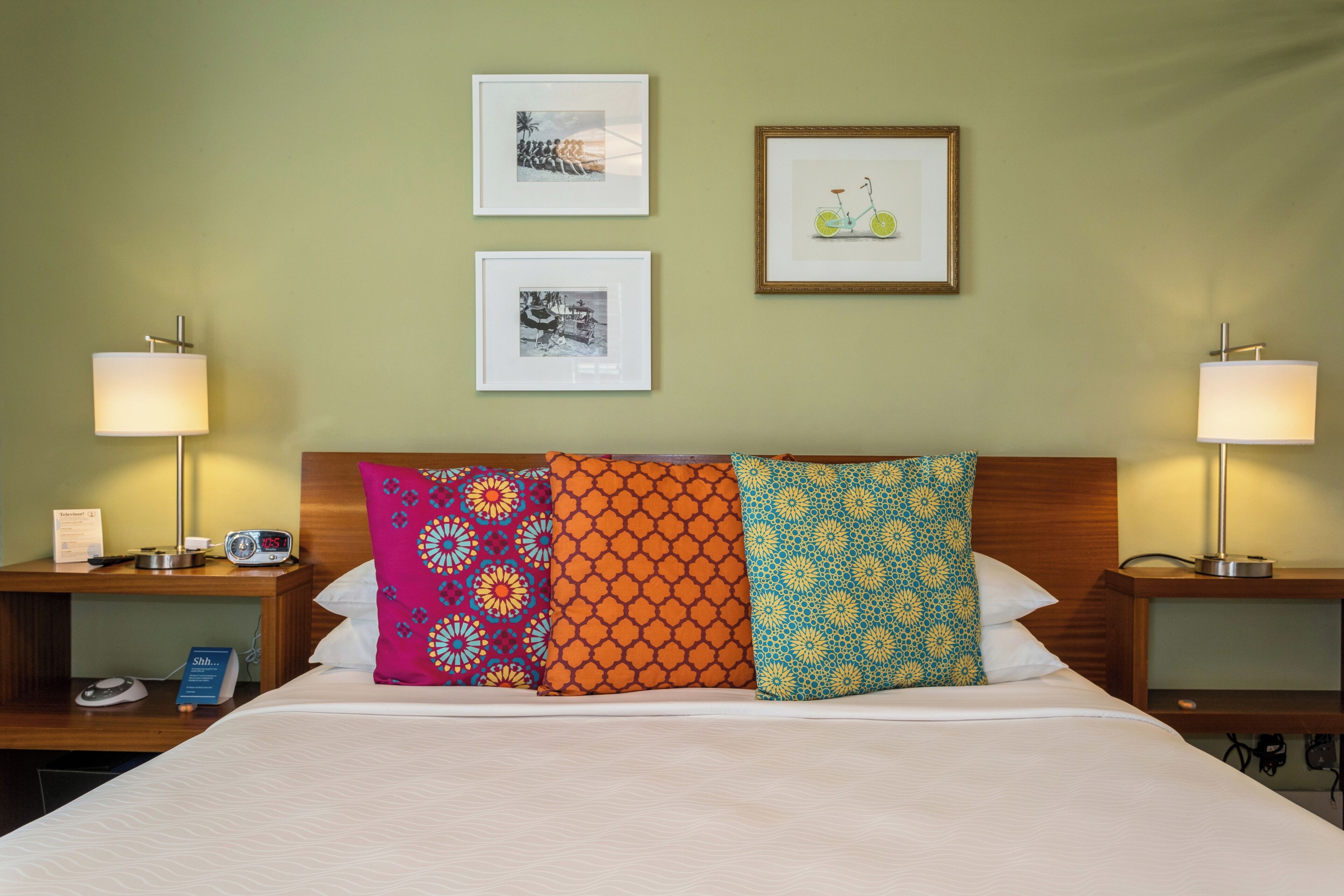 Une chambre d’hôtel avec un lit soigneusement fait, deux tables de chevet avec des lampes et trois tableaux encadrés accrochés au mur.