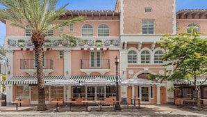 A pink building with a balcony, outdoor seating, and a palm tree.