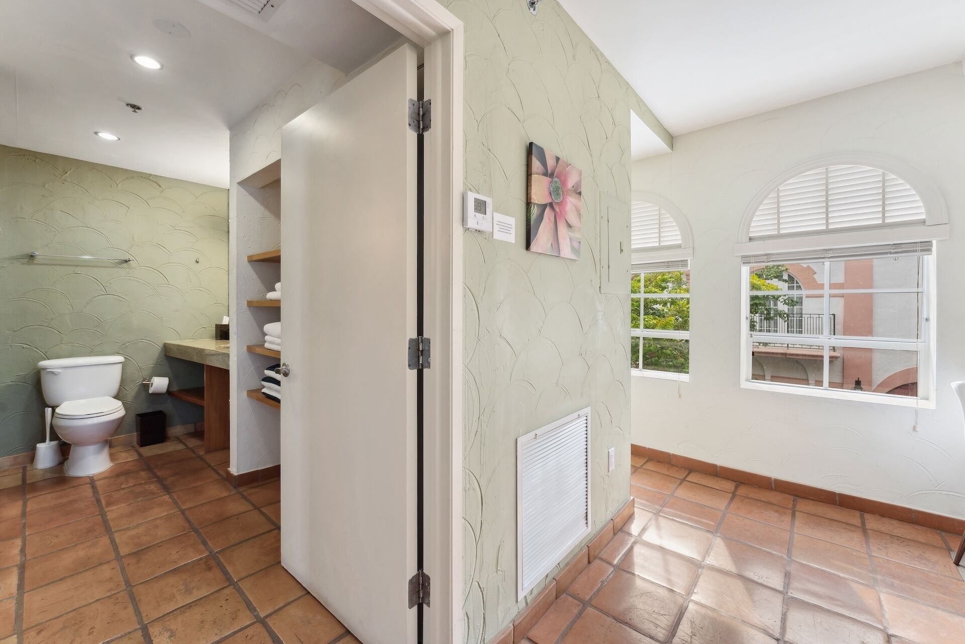 A bathroom with a toilet, sink, and shelves with towels. There are two windows with shutters and a view of greenery outside.