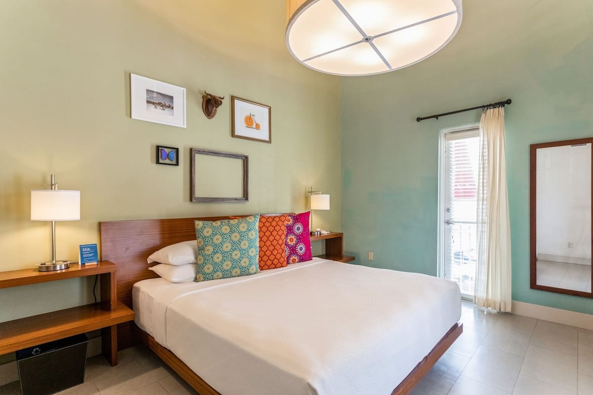 A hotel room with a large bed, two bedside tables, a wall-mounted lamp, a ceiling light, and a window with curtains.