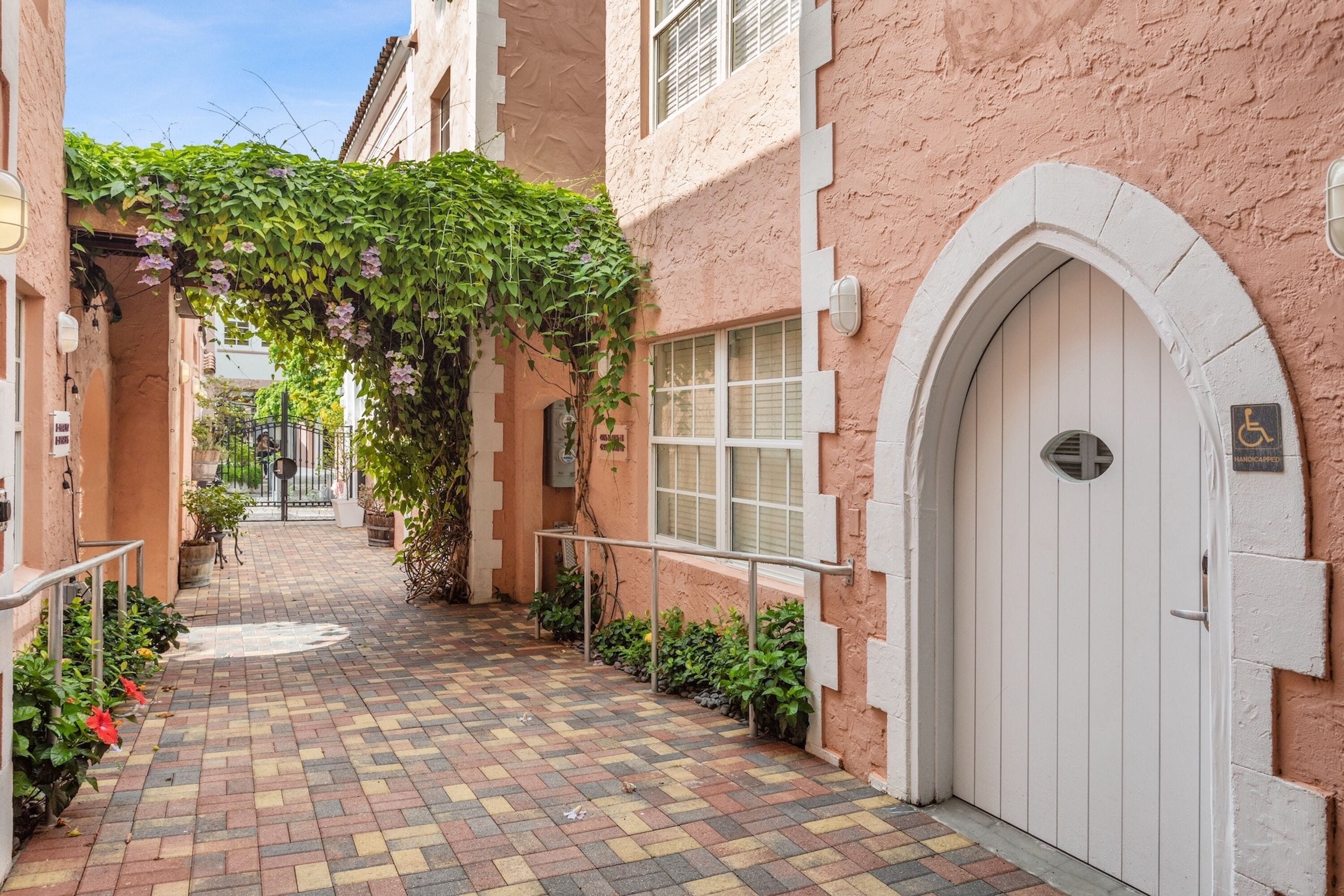 A narrow alleyway with a checkered brick floor, a white door with a wheelchair accessibility sign, and a building with pink walls and a green vine-covered archway.
