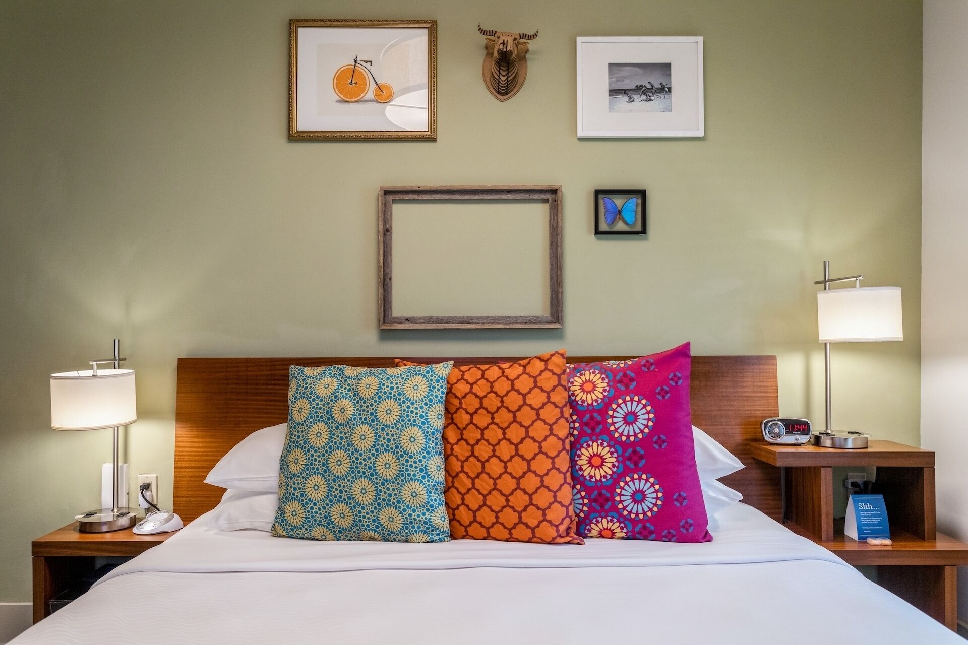 Ein Hotelzimmer mit einem Bett, farbenfrohen Kissen, einem Holz-Kopfteil, einem Nachttisch mit Lampe und gerahmten Bildern an der Wand.
