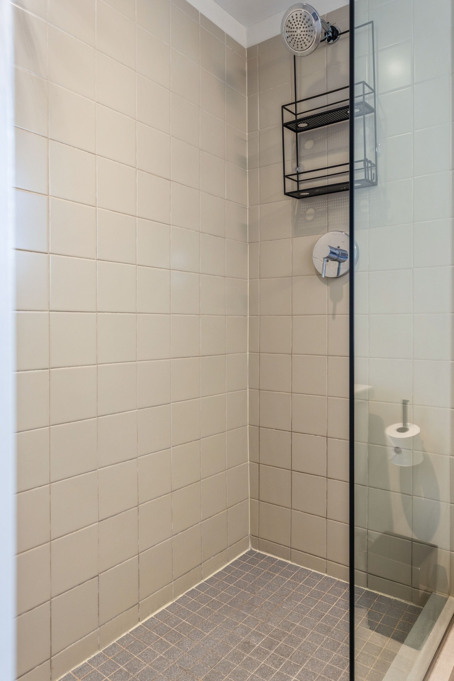 Een moderne badkamer met beige tegels, een glazen douchecabine en een metalen plankunit.