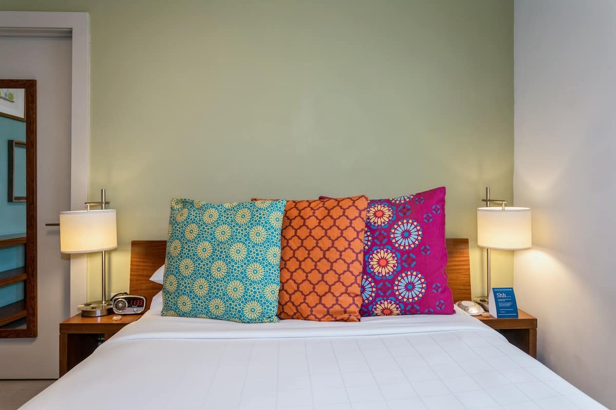 A hotel room with a bed, two bedside tables, a nightstand, a lamp, a clock, and a sign.