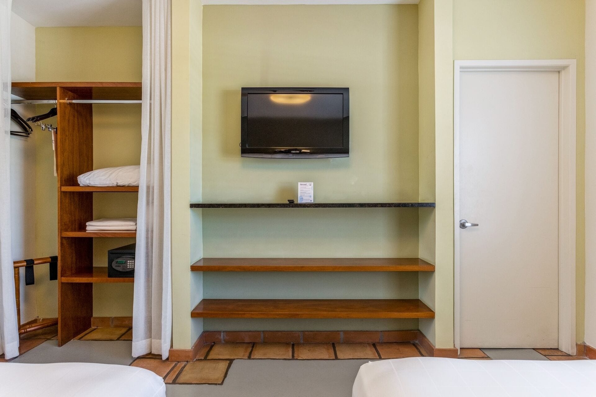 Una habitación de hotel con televisión instalada en la pared, estanterías de madera, una puerta y una cama.