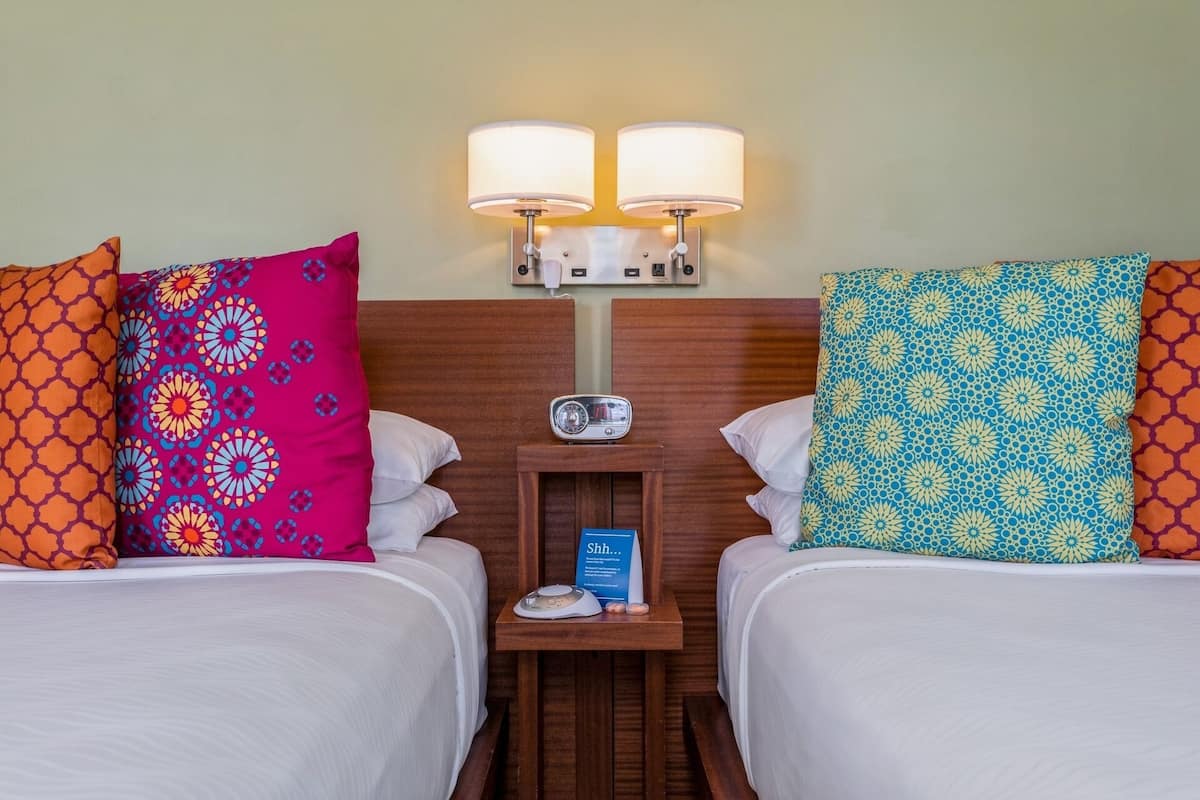 A hotel room with two beds, each with patterned pillows, a bedside table with a radio, and wall-mounted lamps.