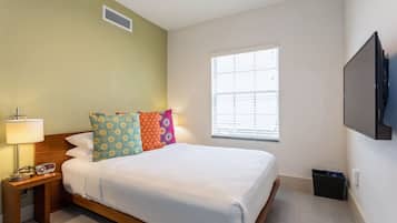 Ein Hotelzimmer mit Bett, Nachttisch mit Lampe und Radio, Fenster mit Jalousien und einem an der Wand montierten Fernseher.