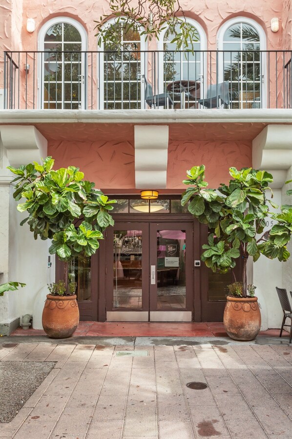A building facade with large windows, a balcony with outdoor seating, and potted plants at the entrance.