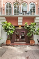 A building facade with large windows, a balcony with outdoor seating, and potted plants at the entrance.
