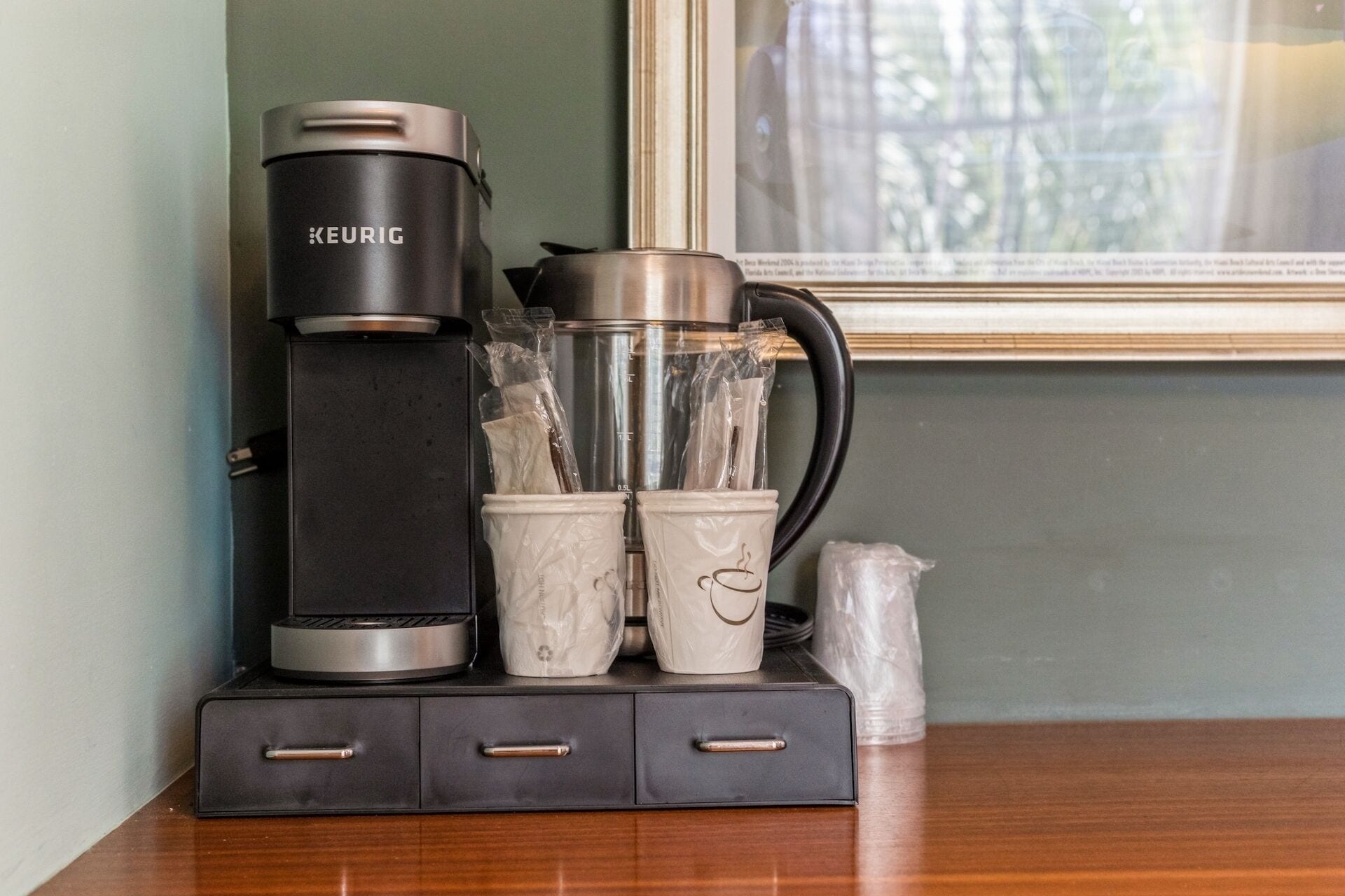 Eine Kaffeemaschine von Keurig, eine Glas-Kanne und zwei Reisebecher auf einem Holztisch.