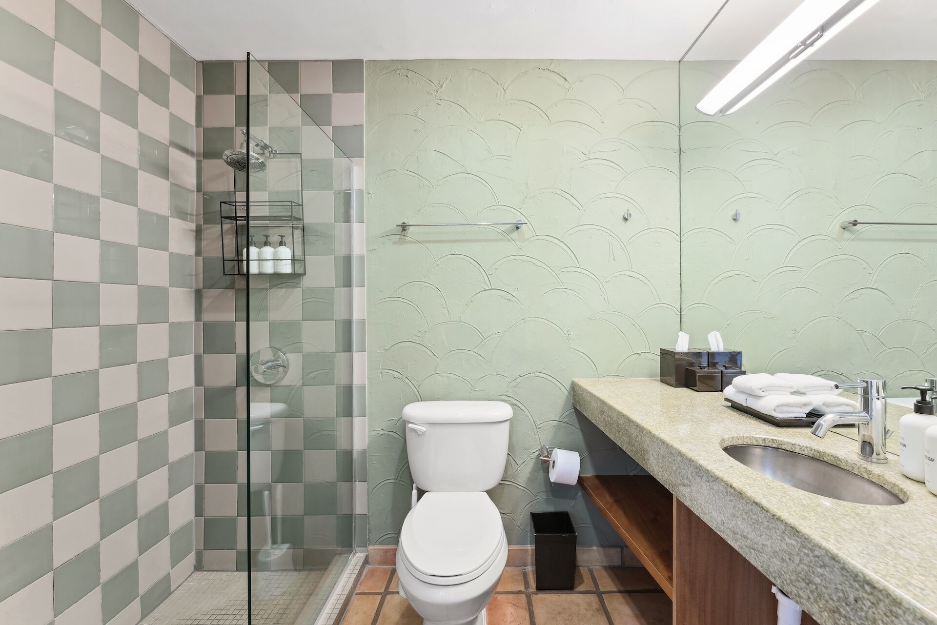 A bathroom with a glass shower enclosure, a toilet, a sink with a towel dispenser, and a mirror.