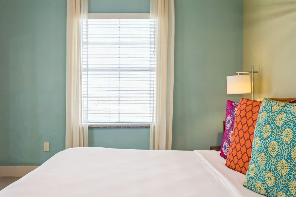 A bedroom with a bed, a window with blinds, and a nightstand with a lamp and colorful pillows.