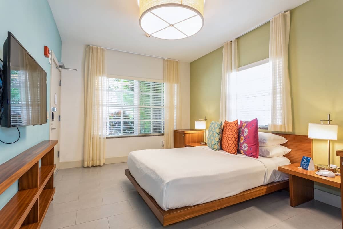 A hotel room with a bed, bedside tables, a TV, a window with curtains, and a ceiling light.