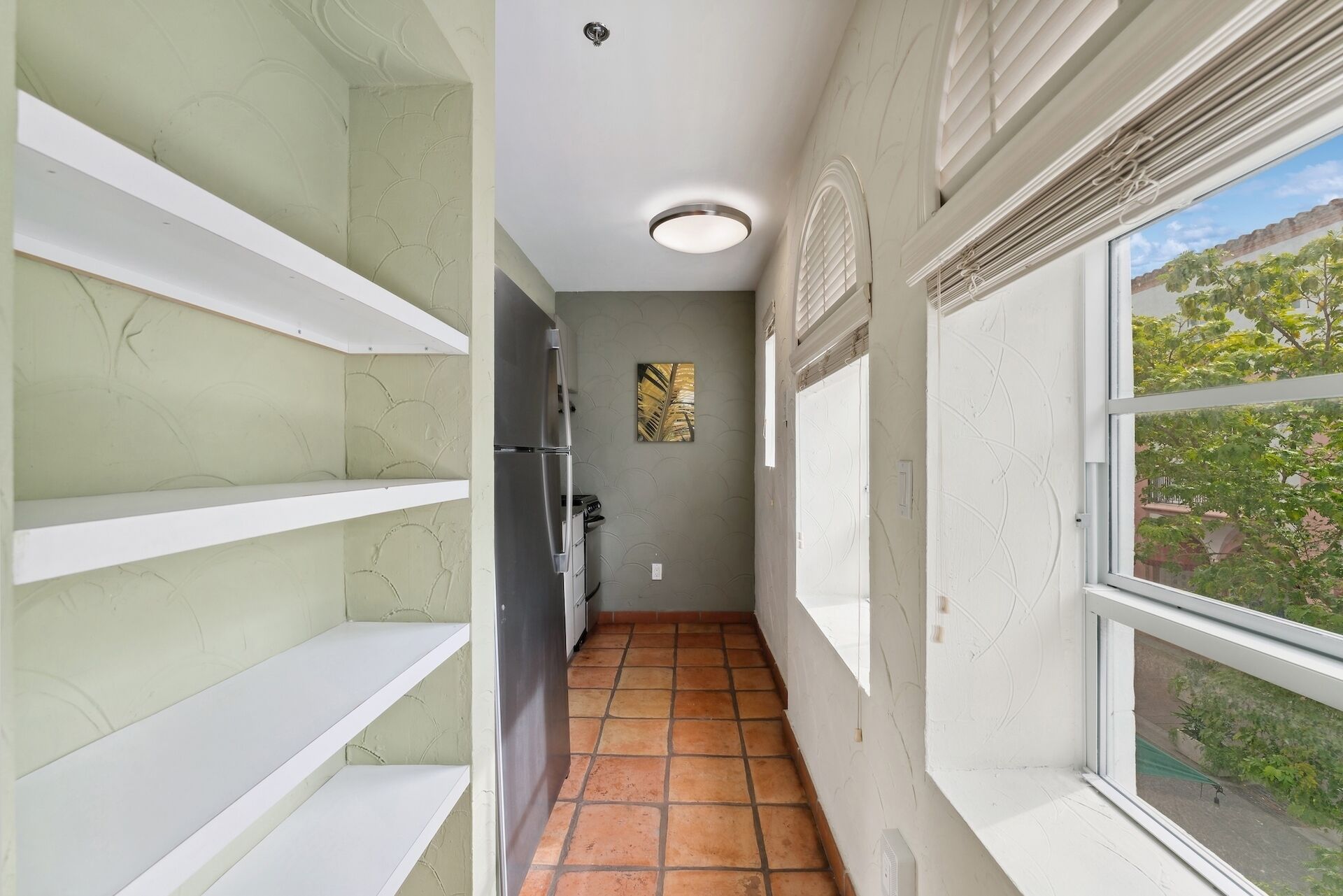 Un pasillo estrecho con estantes blancos, un refrigerador y una ventana que da a zonas verdes.