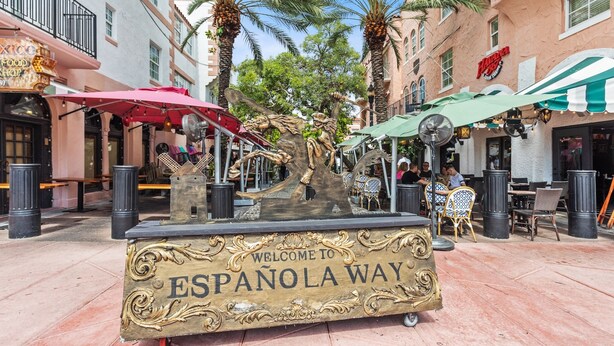 A decorative sign welcoming visitors to ESPANOLA WAY, surrounded by outdoor seating and palm trees.
