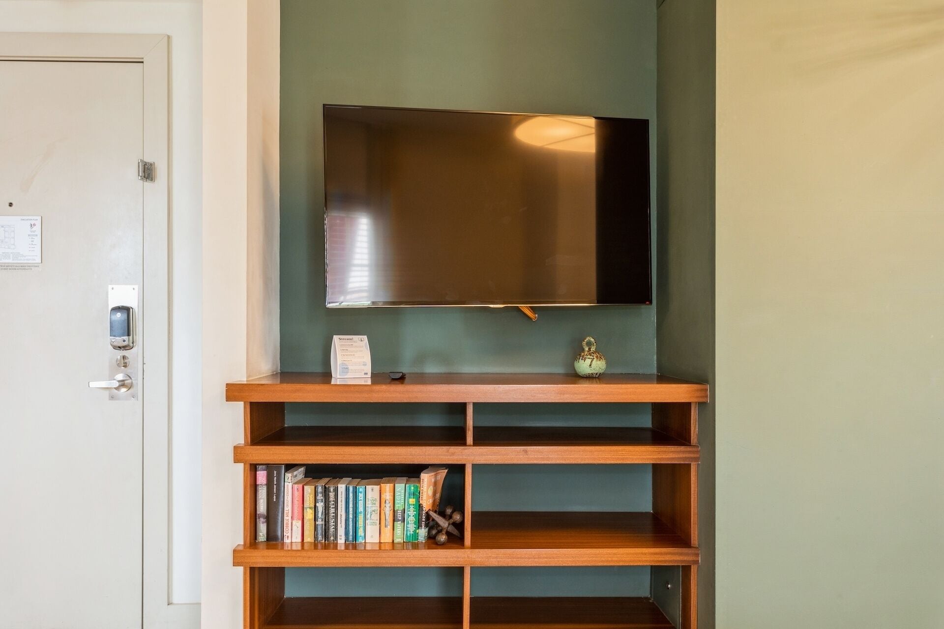 A room with a flat-screen TV mounted on a teal wall, a wooden bookshelf below, and a door with a lock and a keycard reader.