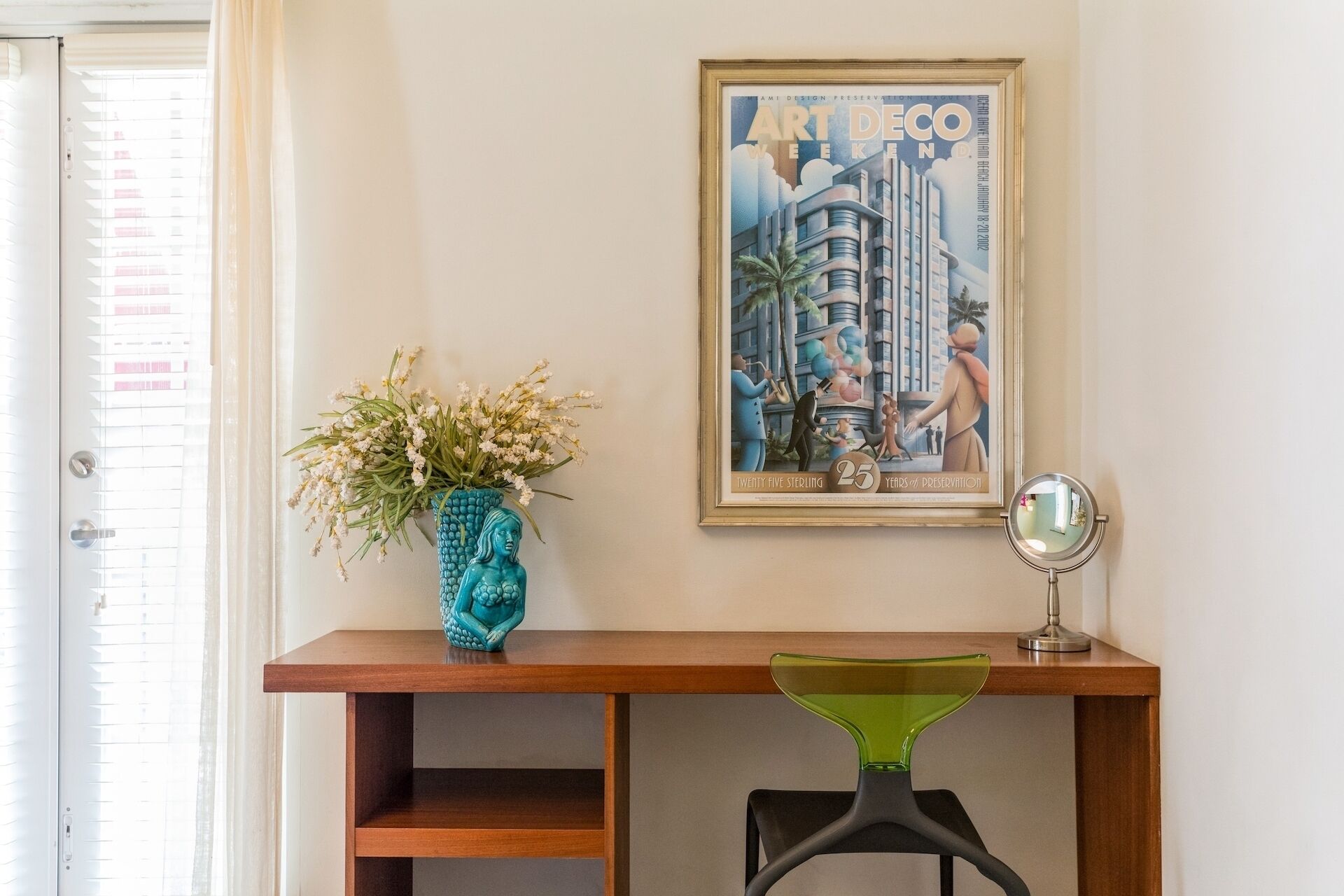 A wooden desk with a framed poster, a vase with flowers, and a green chair.