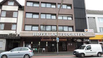 Ein Hotel mit einem Schild, auf dem 'HOTEL AM FLUGHAFFEN' steht, und einem sichtbaren Eingangsbereich, vor dem ein Auto geparkt ist.